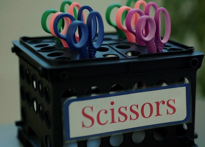 Black storage crate turned upside-down with scissors inserted into the holes, labeled Scissors as an example of dollar store hacks for the classroom 