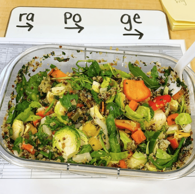 Glass container of salad made from brussels sprouts, pea pods, tomatoes, couscous, and more, sitting on a whiteboard with letters drawn on it