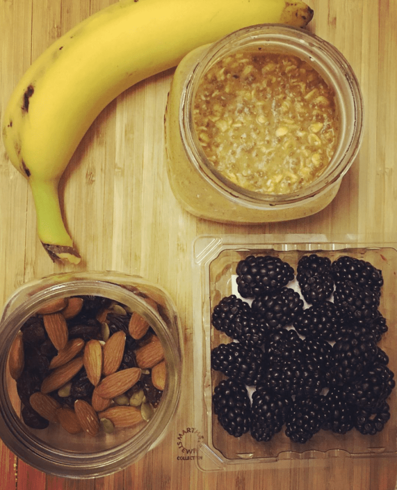 Small glass jars holding overnight oats and mixed nuts, a container of blackberries, and a banana