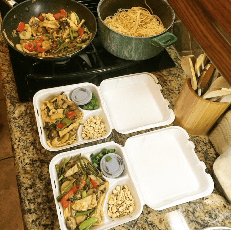 Two pans on a stove, filled with noodles and a stir fry, next to two styrofoam containers of stir fry and side dishes