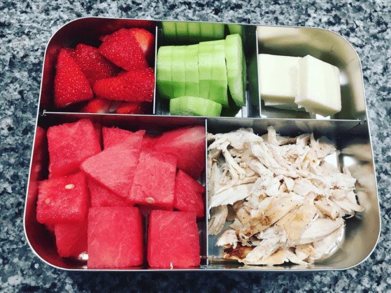 Divided container filled with rotisserie chicken, watermelon, strawberries, cucumber slices, and cheese slices, for a healthy teacher lunch