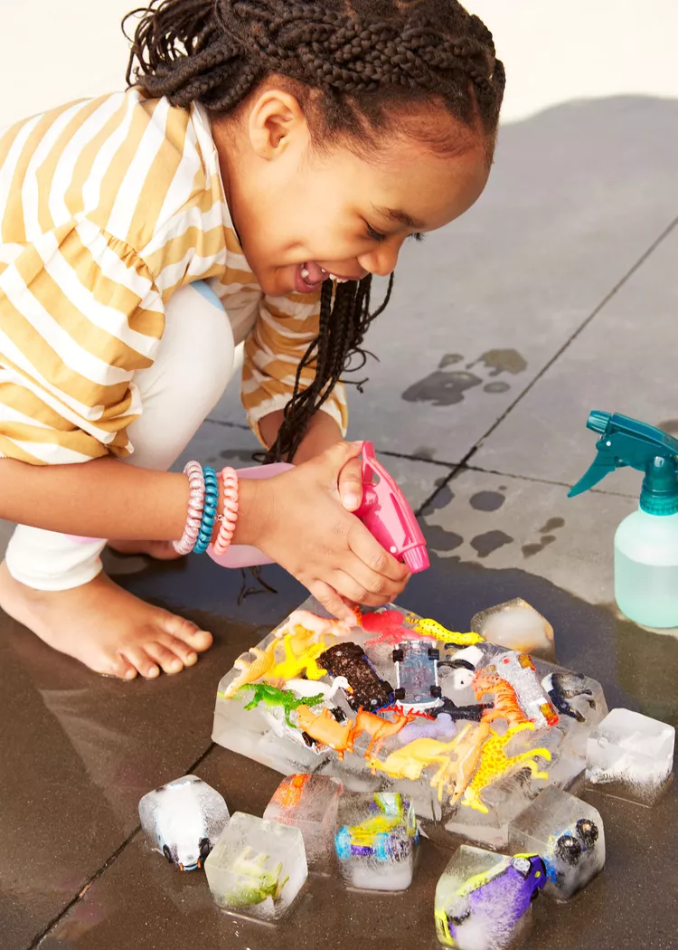 A little ogirl is seen spraying water on a block of ice with small plastic toys frozen into it.